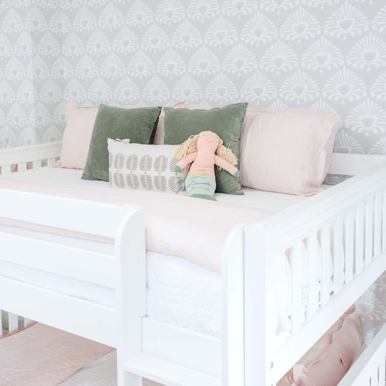 Children's white bunk bed with pastel pink and green bedding, featuring a cute knitted mermaid doll, against a stylish wallpaper background.