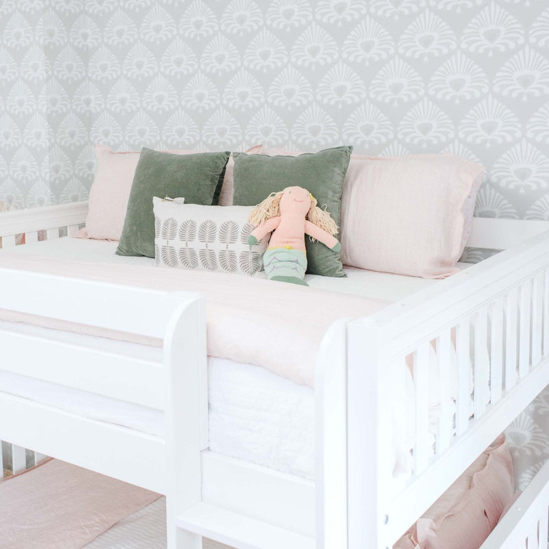 White baby crib with blush pink bedding, green velvet pillows and a handmade doll against light gray patterned nursery wallpaper