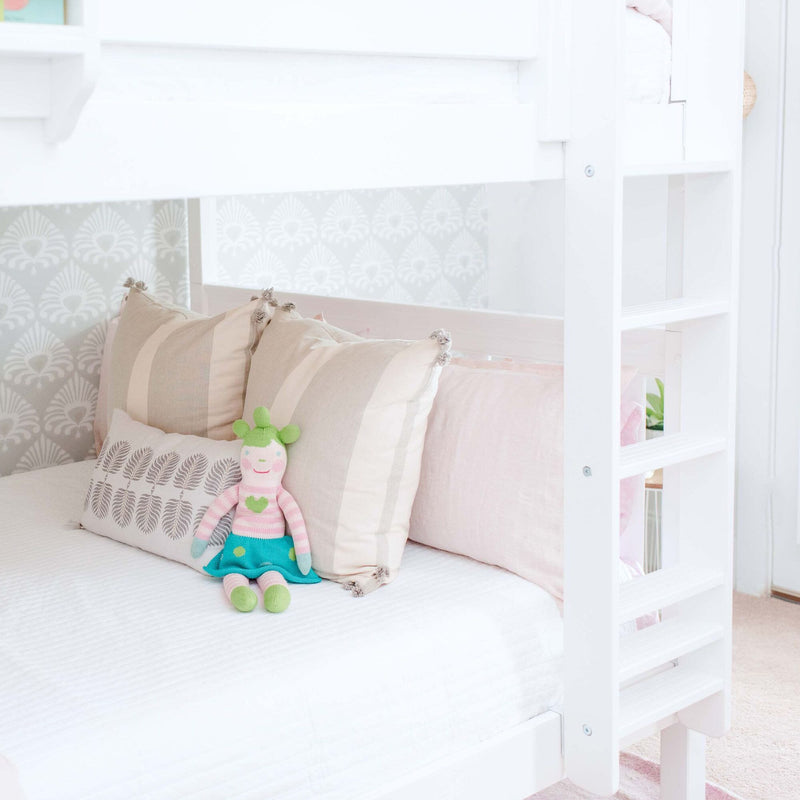 White wooden bunk bed with ladder, pastel pink and beige pillows, patterned wallpaper and knitted doll in a bright children's room