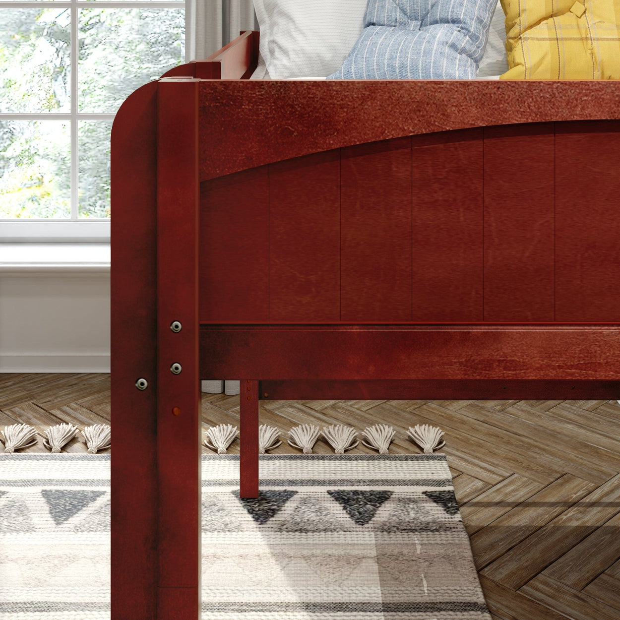 Red wooden bed frame close-up in cozy bedroom interior with decorative pillows, patterned area rug, herringbone hardwood floor and sunlit window