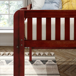 Red wooden baby crib with white mattress and blue and yellow pillows in a sunlit modern nursery with patterned rug and hardwood floor