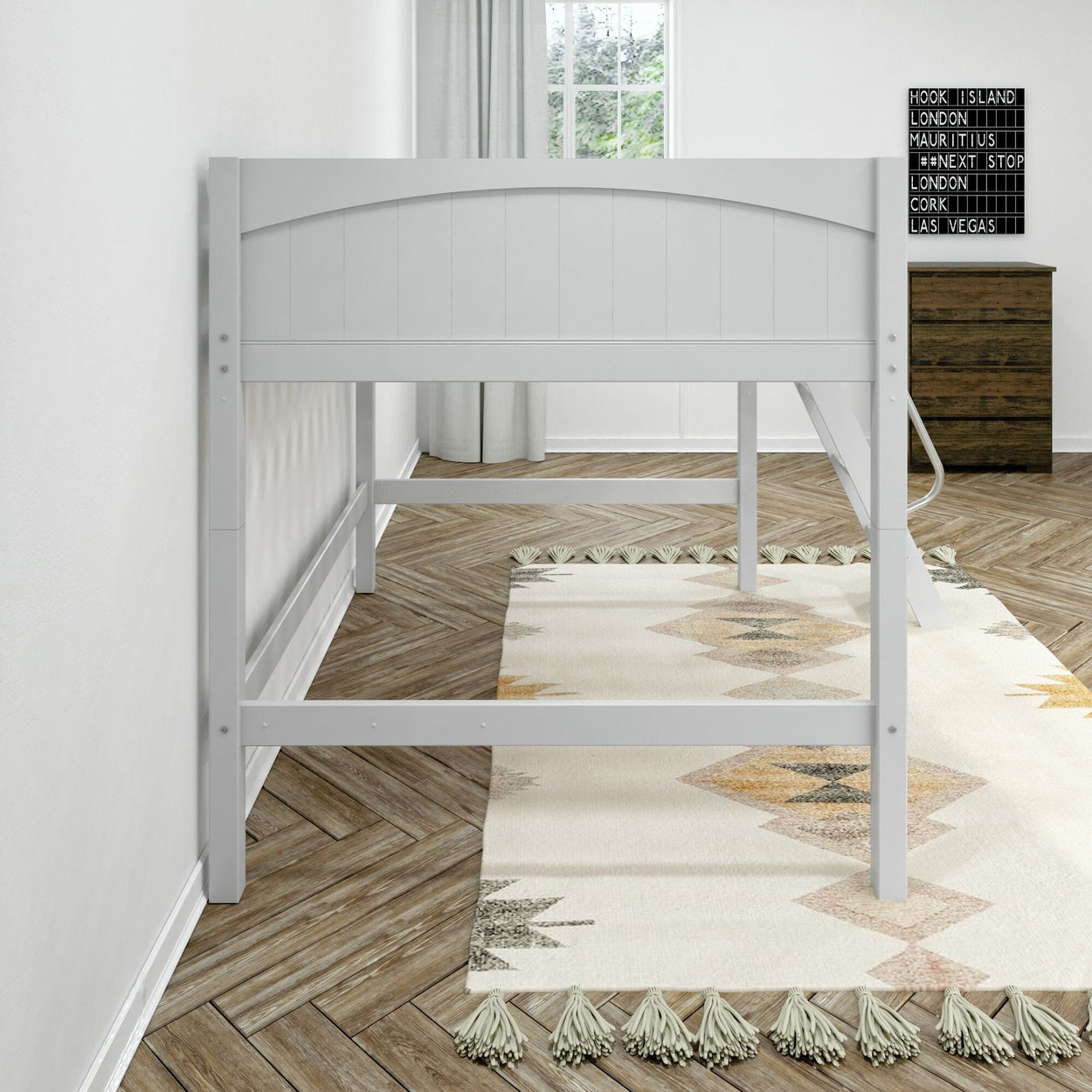 White wooden loft bed frame in a bright minimalist bedroom with geometric tasseled area rug, herringbone hardwood floor, ladder and wooden dresser