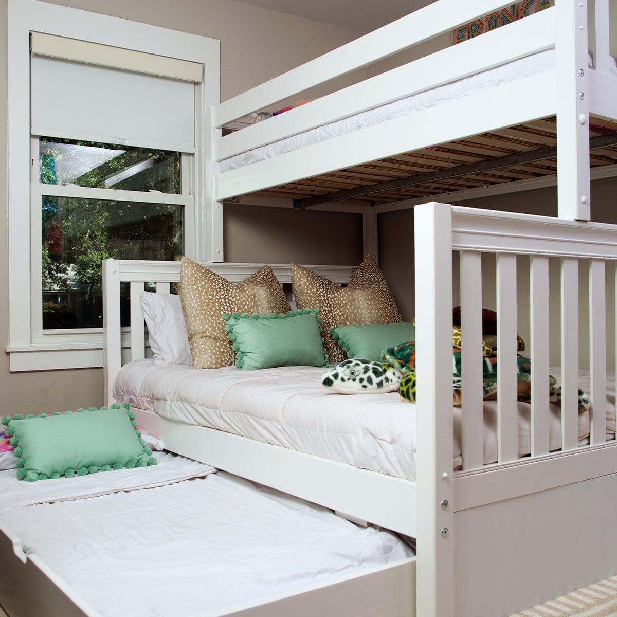 White wooden bunk bed with pull-out trundle in a children's bedroom, white bedding, mint-green pom-pom pillows and tan patterned accent cushions beneath a window