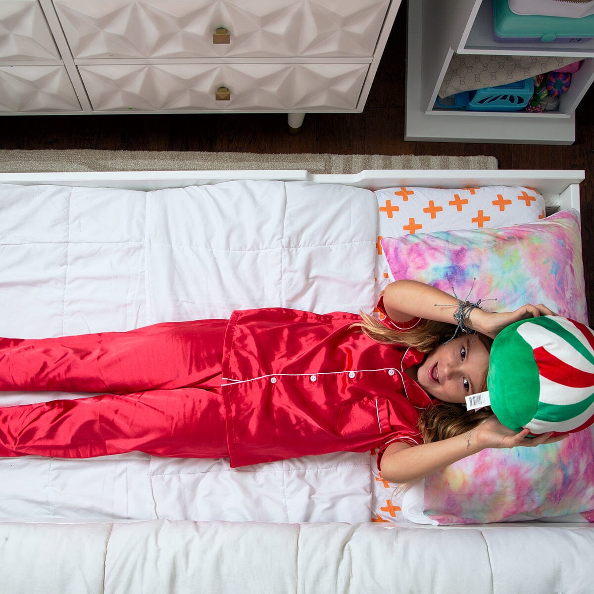 Girl in red satin pajamas lying on a white bed holding a green-and-white plush ball with a tie-dye pillow and kids bedroom decor, cozy children's bedtime and sleepwear