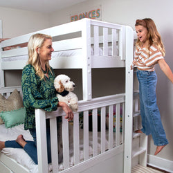Woman and young girl playing on a white wooden bunk bed in a cozy children's bedroom with a small white dog on the lower bunk, ladder, colorful pillows and soft bedding.