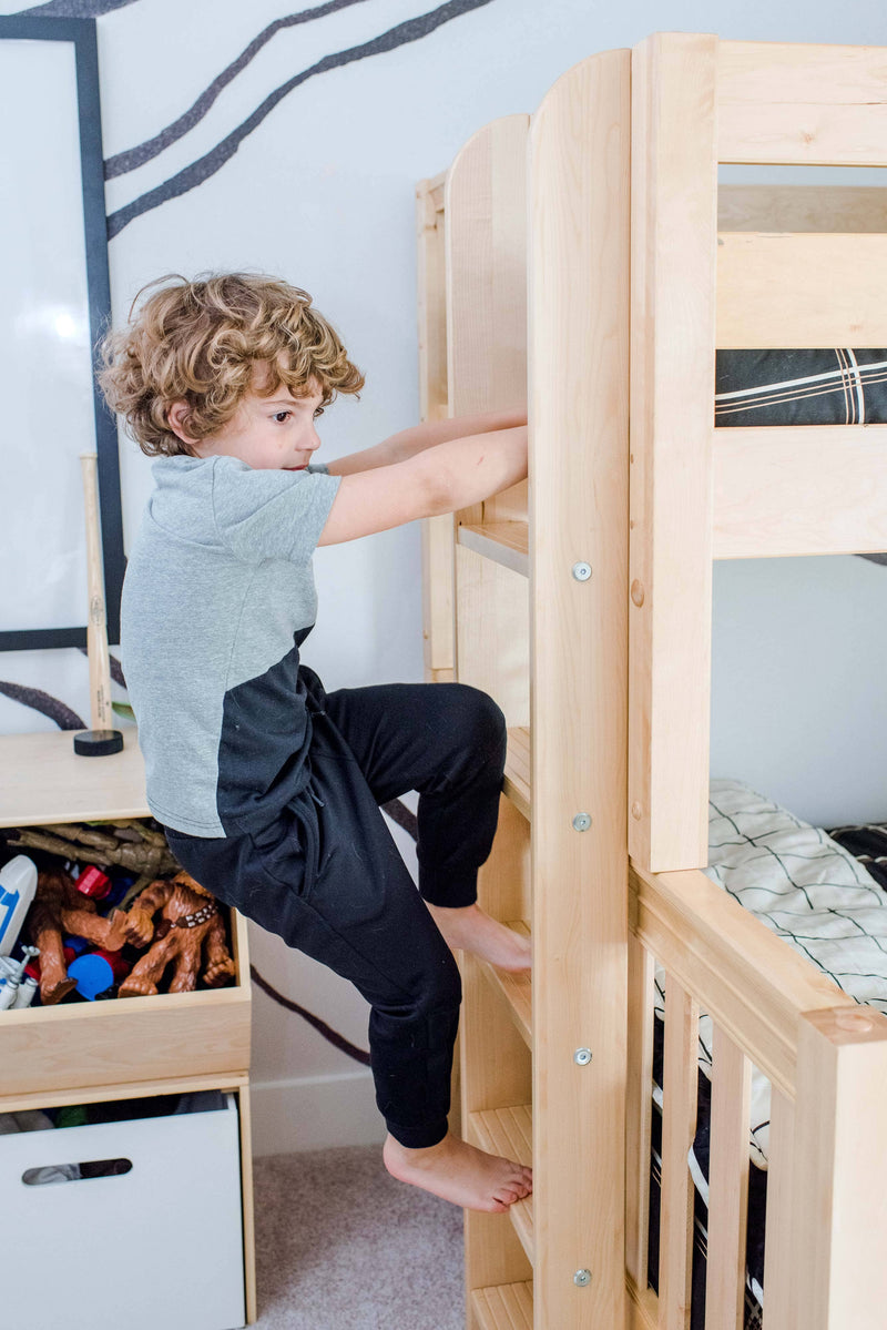Curly-haired young boy climbing a natural wood bunk bed ladder in a bright modern kids' bedroom
