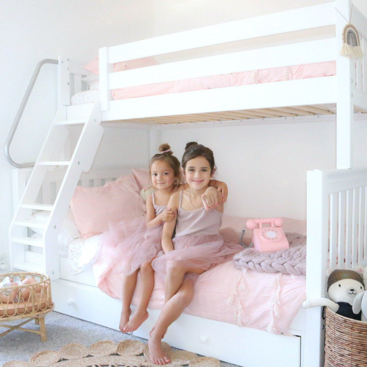 Girls in pink dresses sitting on a white bunk bed in a cozy children's bedroom with pastel decor.