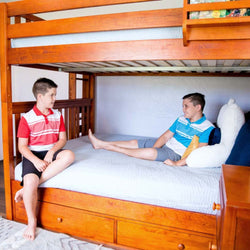 Two boys sitting on a wooden bunk bed with light blue bedding in a bright bedroom.