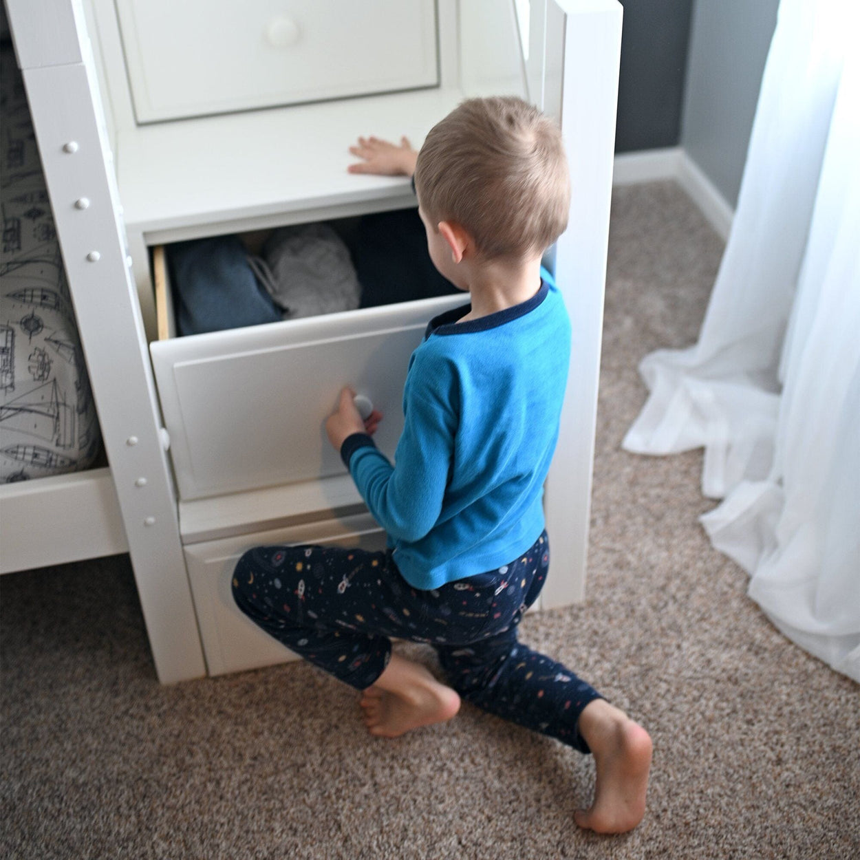 Young child in blue pajamas organizing clothes in a white dresser drawer in a cozy carpeted bedroom.