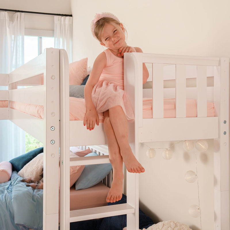 Smiling little girl in pink dress and pearl headband sitting on a white bunk bed in a pastel kids bedroom with pink bedding and string lights