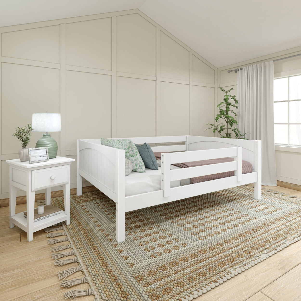 White toddler bed in cozy bedroom with decorative rug, bedside table, and potted plant next to a window.