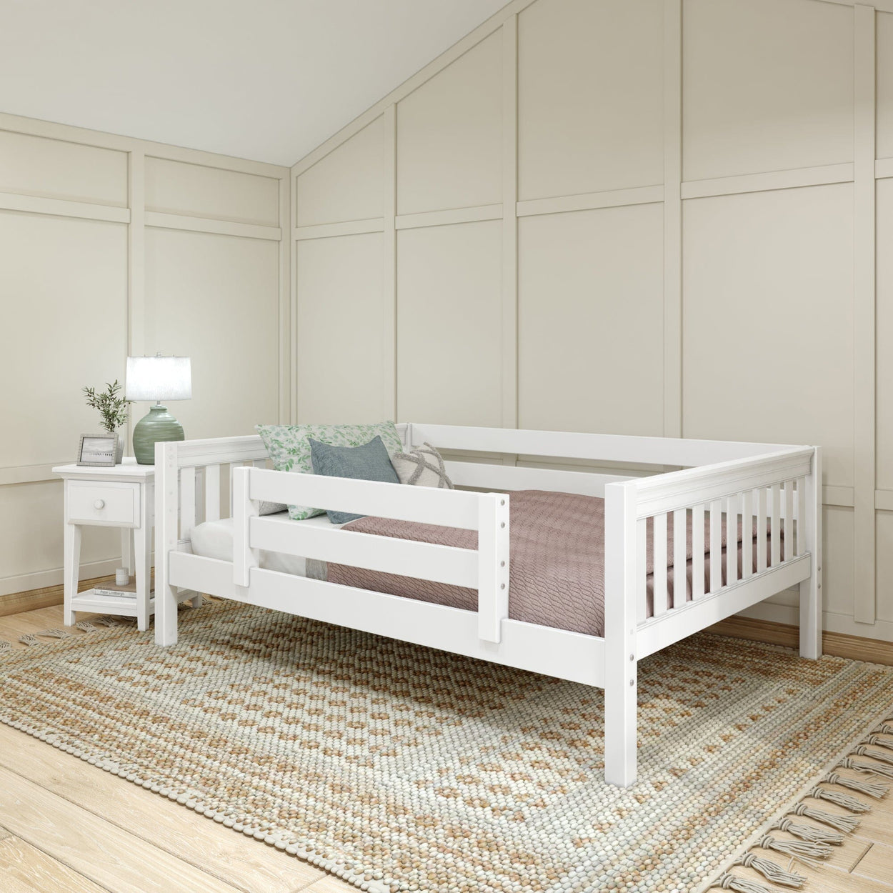 White toddler bed with pink bedding in a cozy room with a bedside table and decorative pillows.