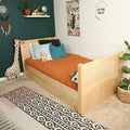 Kids bedroom wooden daybed with under-bed storage drawers, rust-orange bedding and teal pillows, green accent wall, plush giraffe toy and patterned boho rug