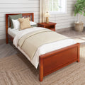 Cozy farmhouse bedroom with wooden single bed, beige layered bedding and knit accent pillows, cherry wood nightstand with pleated lamp, white shiplap wall, woven jute rug and potted plant by a sunlit window
