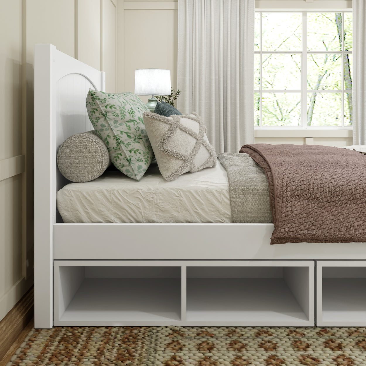 Modern bedroom with white wooden bed frame, plush decorative pillows, and cozy brown blanket by a sunlit window.