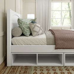 Modern bedroom with white wooden bed frame, plush decorative pillows, and cozy brown blanket by a sunlit window.