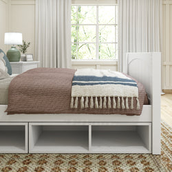 Cozy bedroom interior with a stylish brown bedspread and decorative throw on a bed with storage shelves, beside a white nightstand with a lamp and framed picture.