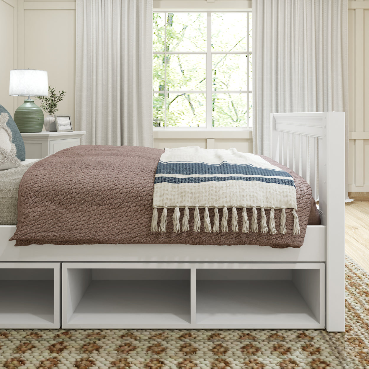 Cozy bedroom with white wooden bed frame, beige patterned blanket, blue and white throw, modern nightstand with lamp, and large window with natural light.