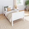 Coastal farmhouse bedroom with white wooden twin bed, neutral layered bedding, green accent pillow, jute rug, shiplap wall, rattan basket and bedside lamp by sunlit window