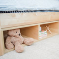 Stuffed teddy bear and books on wooden shelf under a bed with striped bedding.