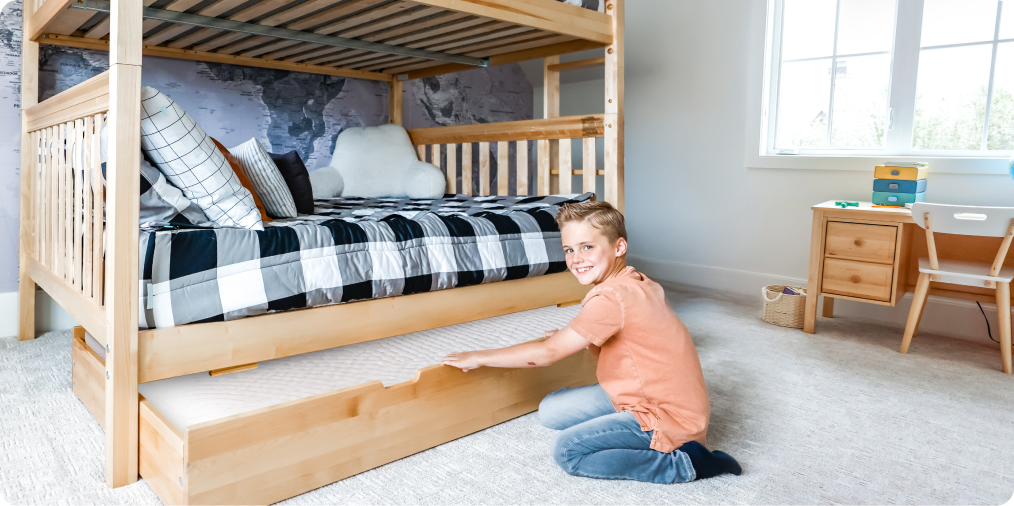Natural wood bunk bed with under-bed trundle being pulled out by a smiling boy in a bright modern kids' bedroom — space-saving storage solution.