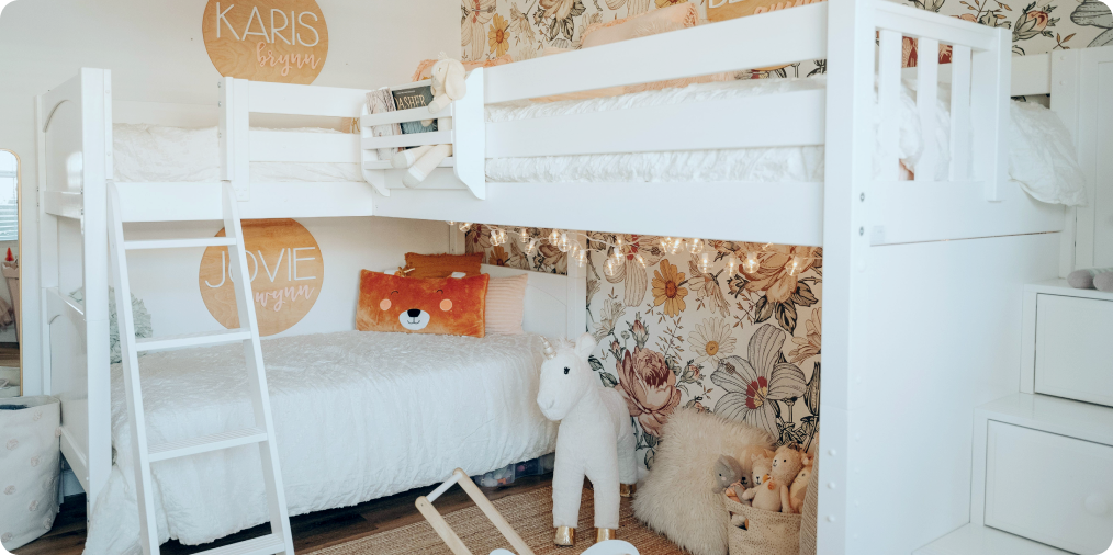White wooden bunk beds in a bright children's bedroom with floral wallpaper, string lights, plush unicorn and bear toys, and personalized name signs.