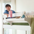 Smiling boy leaning over a white bunk bed in a bright kids' bedroom with a pink tumbler, stacked books and a stuffed toy