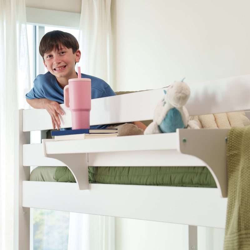 Smiling boy leaning over a white bunk bed in a bright kids' bedroom with a pink tumbler, stacked books and a stuffed toy