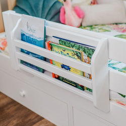 White wooden bed-rail book pocket holding colorful children's board books on a toddler bed – bedside book organizer for kids' bedroom reading nook