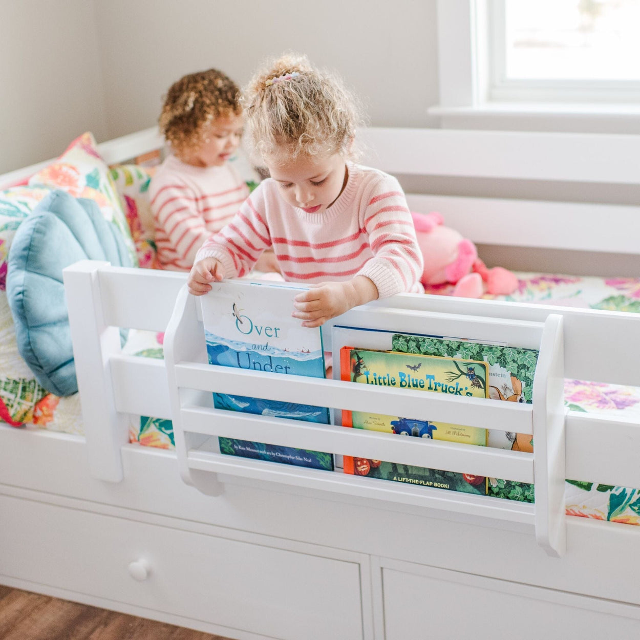 Two young girls reading children's books on a white daybed with attached bed-rail bookshelf and colorful kids room decor