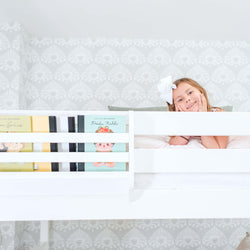 Smiling young girl lying on a white bunk bed with built-in bookshelf and children's books in a modern gray patterned kids' bedroom