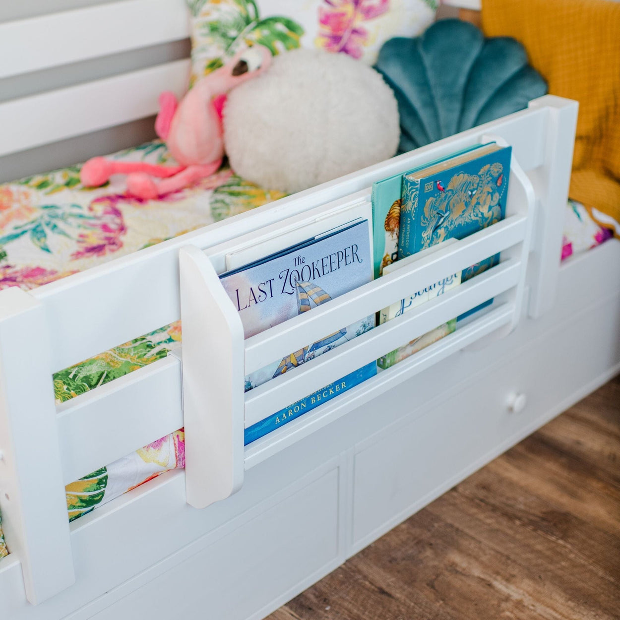 White kids bed with attached bedside book pocket holding children's picture books, colorful floral bedding and plush toys