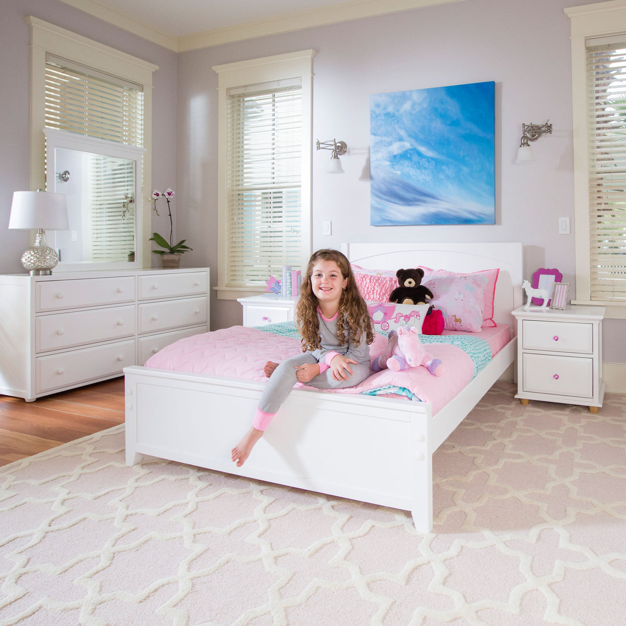 Child smiling in a pastel-themed bedroom with white furniture, pink bedding, and plush toys.