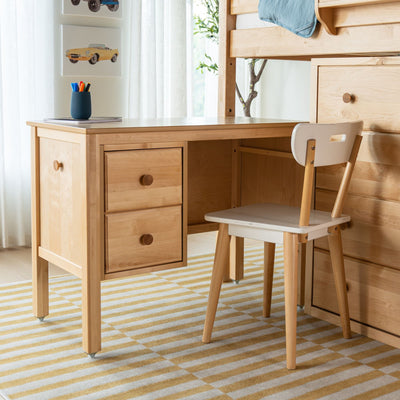 Natural wood kids' study desk with two drawers and matching chair under a loft bed in a bright modern children's bedroom, striped rug and pencil holder