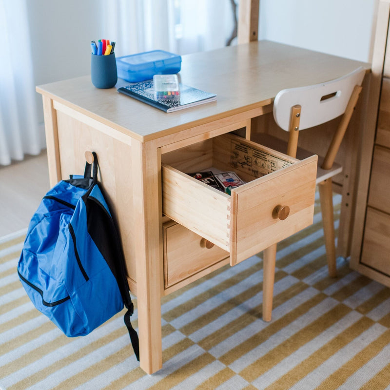 Wooden kids study desk with open drawer, blue backpack hanging at side, composition notebook and colorful pens on the tabletop