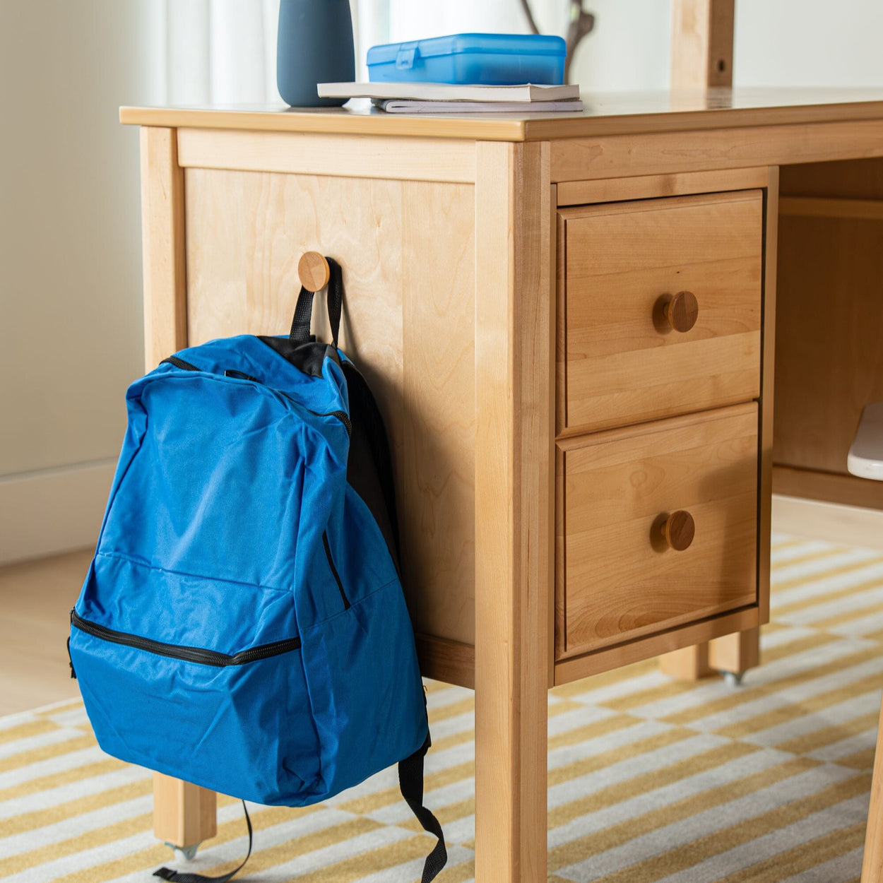 Blue backpack hanging from knob of light wood desk with drawers in bright home study area on striped rug