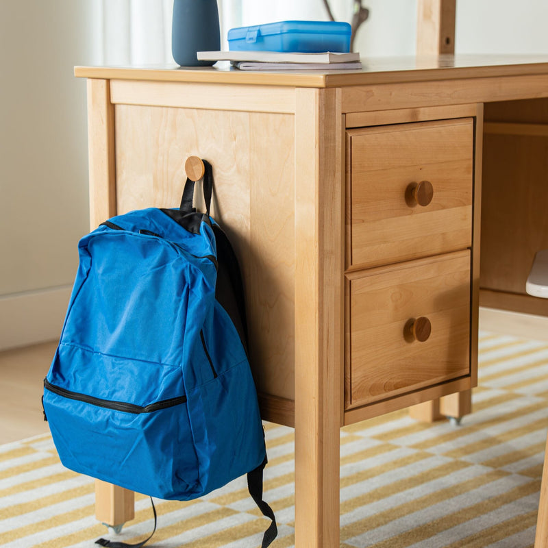 Light wood student desk with three drawers, blue backpack hanging on a knob and blue lunchbox and notebooks on top, home study area on striped rug
