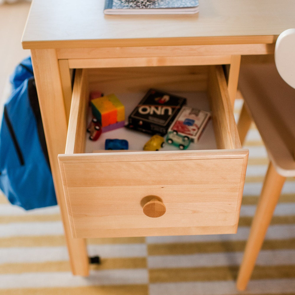 Open children's wooden desk drawer with UNO cards, small toy cars and a colorful puzzle cube; blue backpack and notebook nearby on a striped rug.