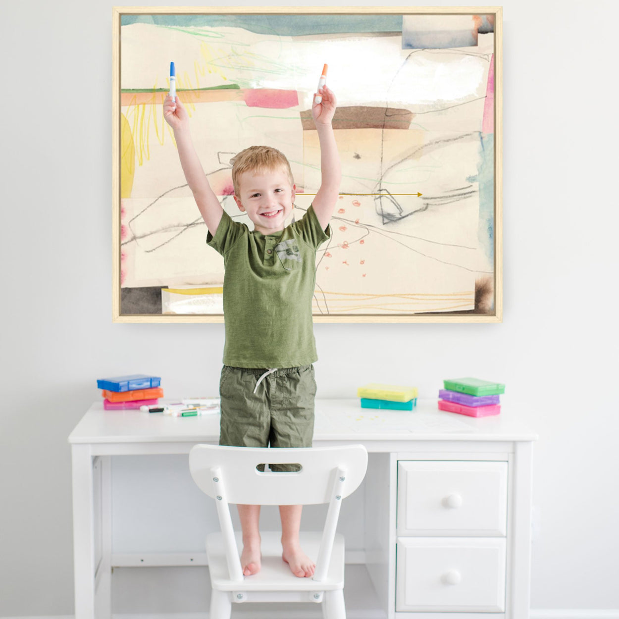 Child standing on chair drawing at white desk with colorful markers and abstract art on wall.