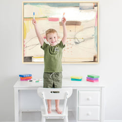 Child standing on chair drawing at white desk with colorful markers and abstract art on wall.