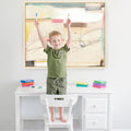 Child standing on chair drawing at white desk with colorful markers and abstract art on wall.