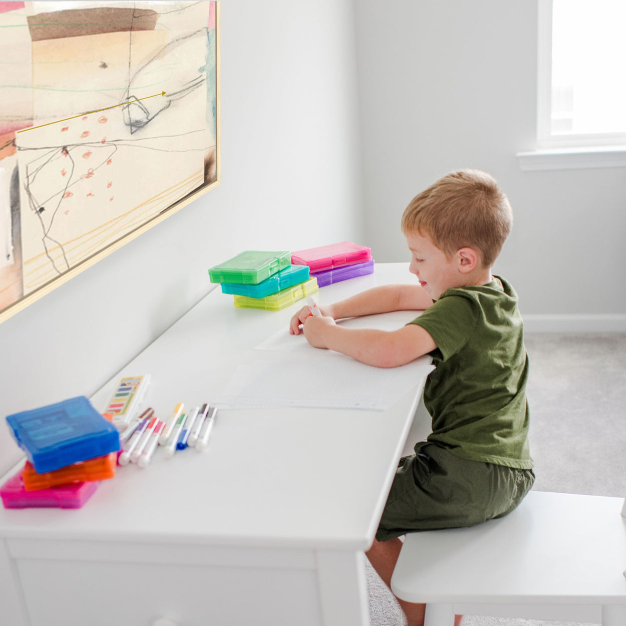 Child sitting at desk drawing with markers, colorful storage boxes on the table, engaging in creative art activity at home.