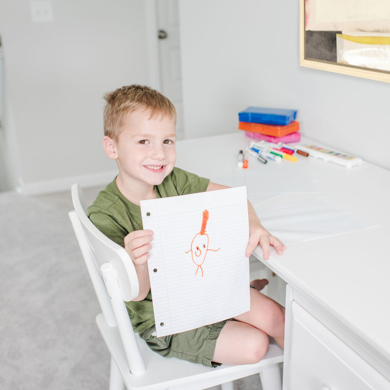 Young boy smiling and showing his drawing at a desk with colorful markers, highlighting creativity and childhood art.