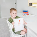 Young boy smiling and showing his drawing at a desk with colorful markers, highlighting creativity and childhood art.