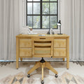 Minimalist light-wood home office setup with four-drawer desk and rolling wooden chair by a black-framed window showing greenery, desk lamp, books, and pencil cup.