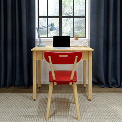 Minimal home office desk setup with wooden desk, red chair, laptop, blue desk lamp, pencil holder and alarm clock by window with navy curtains