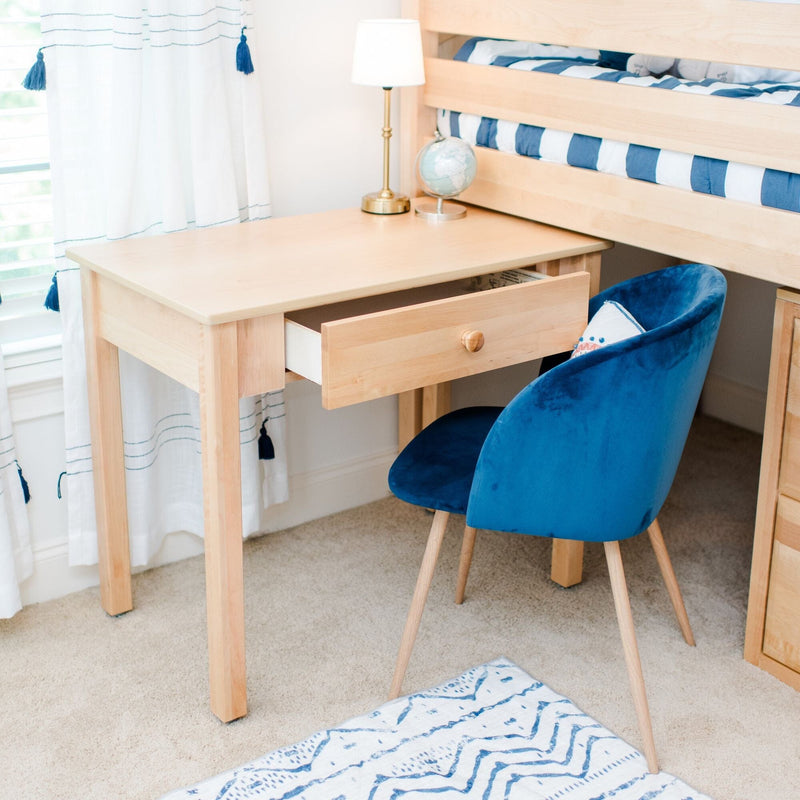 Light wood study desk with open drawer and brass lamp beside a blue velvet chair under a bunk bed in a bright coastal-style kids bedroom