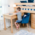 Kids bedroom study nook: child sitting at a natural wood desk beneath a loft bed with blue-striped bedding, blue velvet chair, lamp and study supplies — home study area for kids.