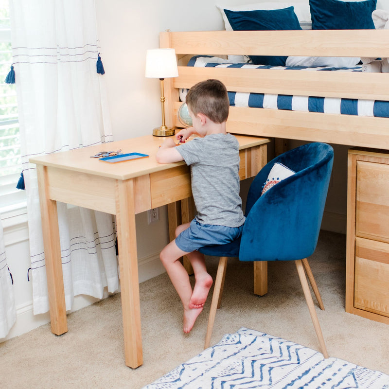 Kids bedroom study nook: child sitting at a natural wood desk beneath a loft bed with blue-striped bedding, blue velvet chair, lamp and study supplies — home study area for kids.