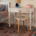White children's desk and chair with wooden legs beside bed, navy table lamp, stacked books and pink makeup pouch on glossy tabletop over colorful boho rug